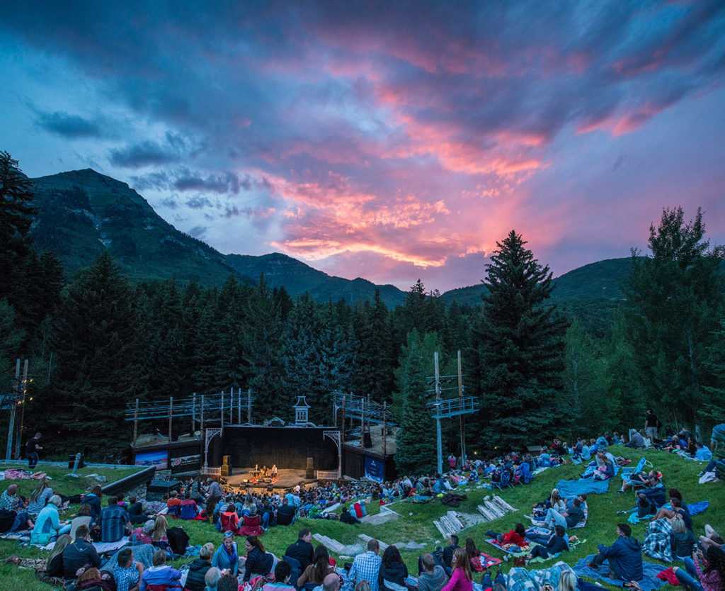 An outdoor theater performance at dusk with a vibrant sunset sky. spectators relax on the grassy hillside, enjoying the scenic mountain backdrop.