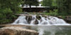 A serene scene featuring a cascading waterfall over rocks in the foreground with a cozy cabin surrounded by lush greenery in the background at Sundance Resort.