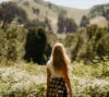 A woman with long blonde hair, seen from behind, stands in a lush field with tall grass and wildflowers, gazing towards a green, tree-covered hill while on one of the hiking trails in