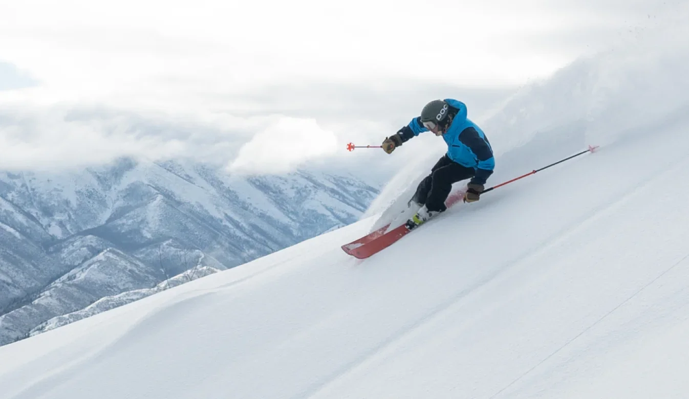 A skier in a blue jacket and black pants carves through fresh powder on a steep, snowy slope, creating a trail of snow. Snow-covered mountains and a cloudy sky form the background, showcasing the ultimate adventure for anyone with a season pass.