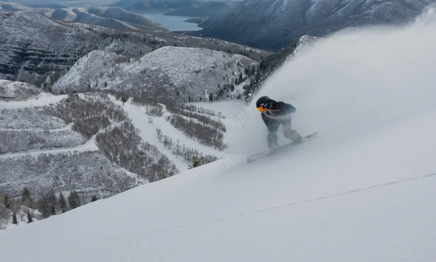 A snowboarder dressed in dark clothing and a helmet carves through fresh powder on a snowy mountain slope, making the most of their season pass. Forested hills and a distant lake are visible in the background under an overcast sky. The snow kicks up around the snowboarder, creating a dynamic action shot.