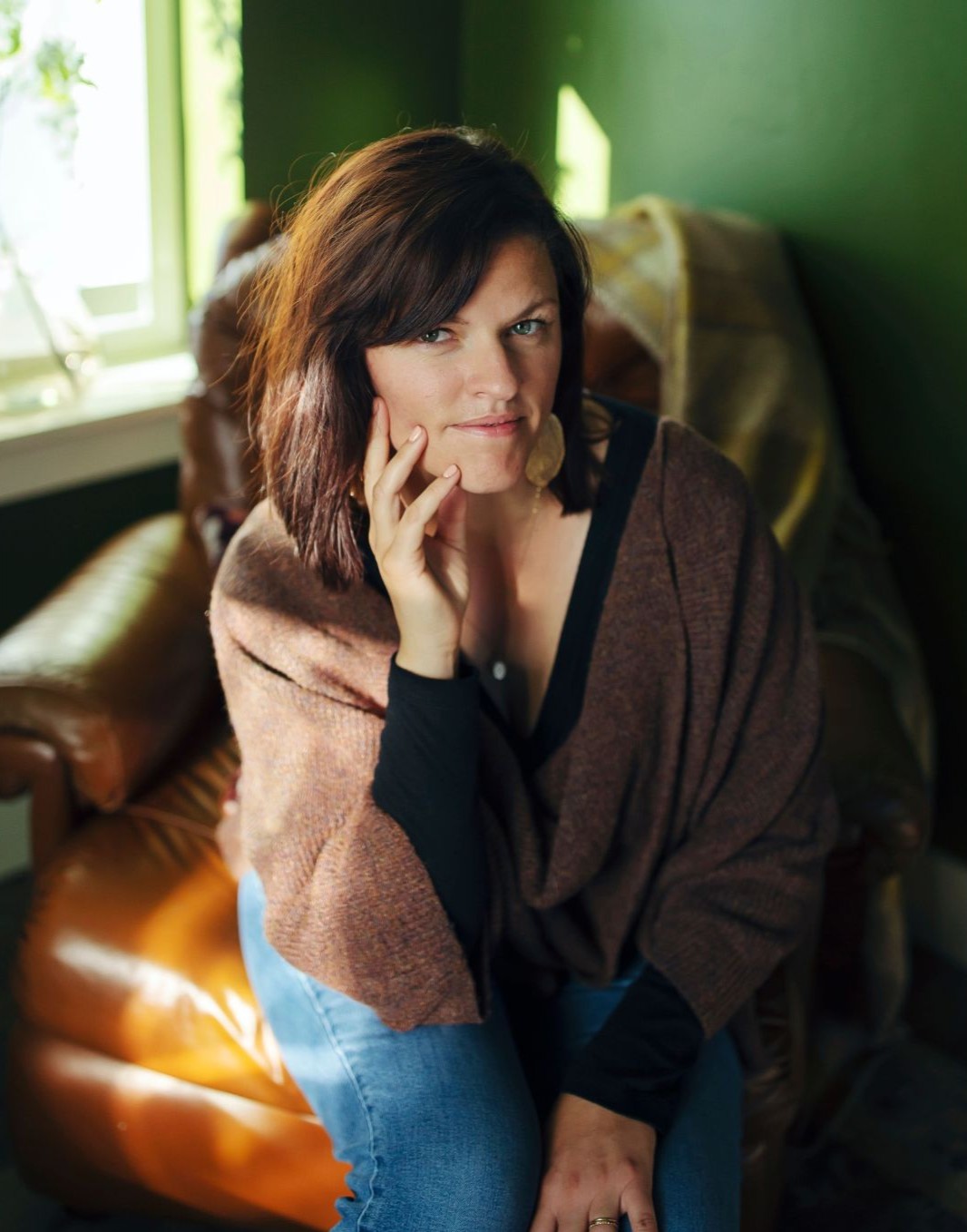 A woman sits in a cozy, sunlit room on a leather chair. She wears a brown shawl over a black top and jeans, with her hand resting on her cheek. The background features green walls and a window with a plant.