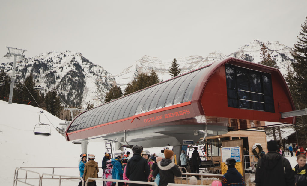 A ski lift station named "Outlaw Express" at Sundance Resort Utah, with skiers in winter gear gathered around, set against a backdrop of snow-covered mountains.