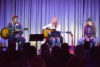 Three musicians perform on stage in front of an audience, two playing acoustic guitars and one singing into a microphone, with soft purple lighting in the background.