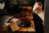 A person's hand is slicing a piece of cooked meat on a wooden cutting board using a knife and fork. The meat appears well-cooked and juicy, with a dark, crispy exterior. An empty card holder is placed in front of the meat.