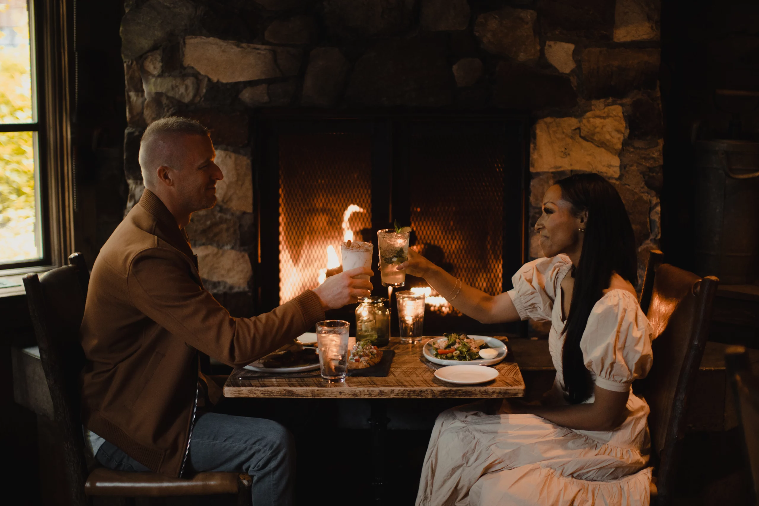 A couple toasting drinks at a cozy, dimly lit dinner by a fireplace, with a rustic stone backdrop. they sit across from each other, smiling in an intimate setting.