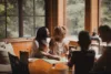 A family engaged in a board game in a cozy, sunlit room with large windows overlooking a forest. a mother smiles at her daughter, while a young boy and another adult focus on the game.