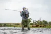 A person dressed in outdoor gear is fly fishing in a shallow river. They have a fishing net attached to their backpack. In the background, a herd of cattle grazes in a grassy area. The sky is overcast.
