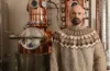 A man wearing a traditional patterned sweater stands confidently in front of a copper distillation apparatus, with elements of the still reflecting the surrounding room.