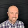 A bald man with a short beard, wearing a purple and white plaid shirt, is smiling at the camera. The background is black.