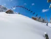 A snowboarder performs a high jump off a wooden ramp in a snowy landscape, captured in multiple frames showing their aerial progression. The scene is set against a backdrop of clear blue sky, snow-covered trees, and a steep snowy slope.