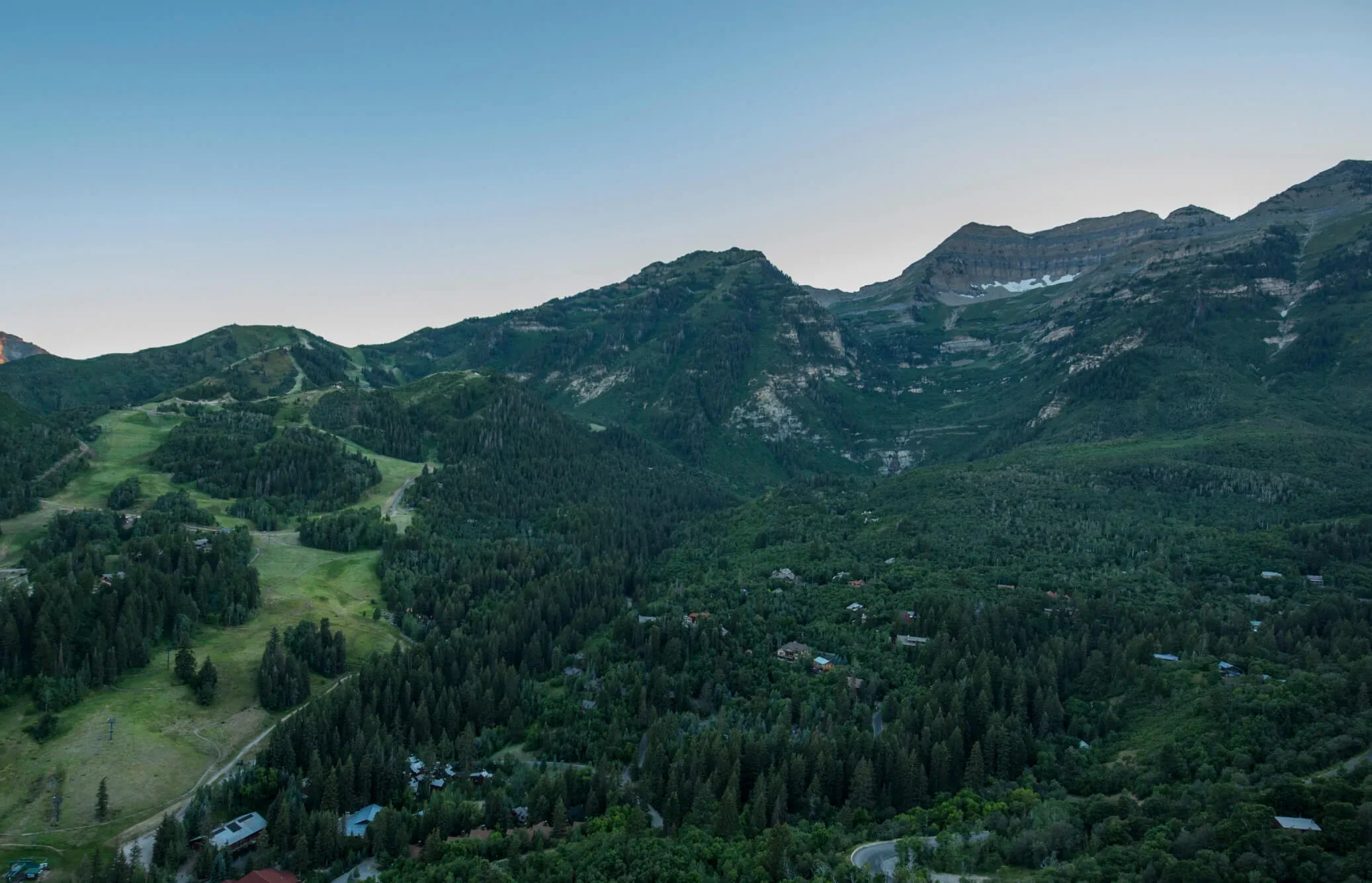 A lush green valley surrounded by forested mountains under a clear sky at dusk. Scattered houses are visible among the trees, and ski slopes and trails wind their way through the landscape, leading up to the mountain peaks.