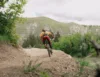 A person wearing a helmet and orange jacket rides a mountain bike on a dirt trail through a lush, green valley with hills in the background. The path is lined with trees and bushes under an overcast sky.