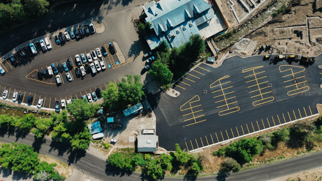 Aerial view of two parking lots separated by a road. The left lot is mostly filled with cars, while the right lot is newly paved and empty. Trees and a few buildings surround the area, creating a mix of natural and urban elements.