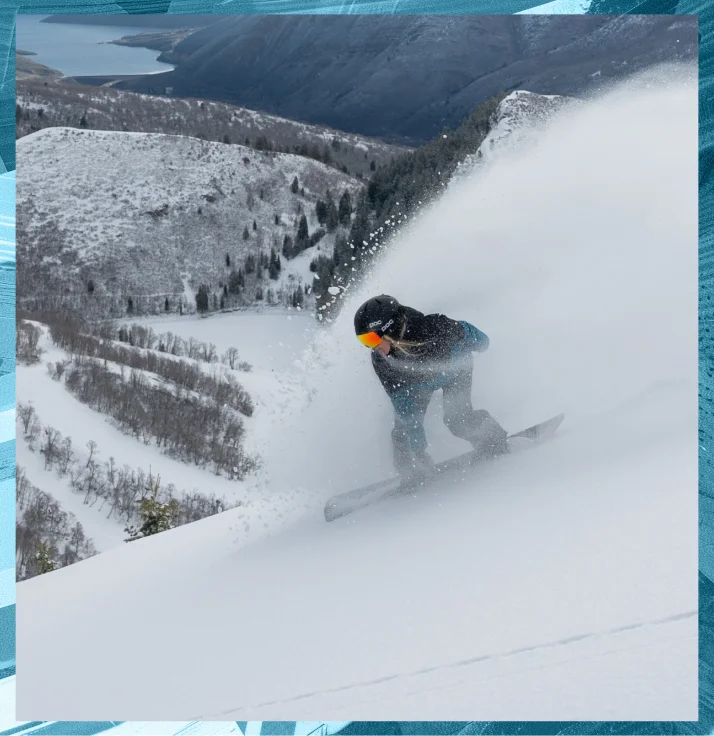 A snowboarder wearing a helmet and goggles is carving through deep powder snow on a steep mountain slope. Snow sprays up around them as they make their turn, reminiscent of an expert maneuver seen in an S-Card video. The background features snow-covered trees and mountain ridges under a cloudy sky.