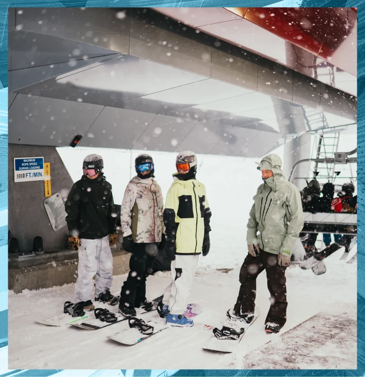 Four snowboarders in winter gear stand in the snow in front of a chairlift station, S-Cards ready. Snow is falling around them, and they are wearing helmets, goggles, and warm jackets. They appear ready to board the chairlift for a run down the snowy slopes.