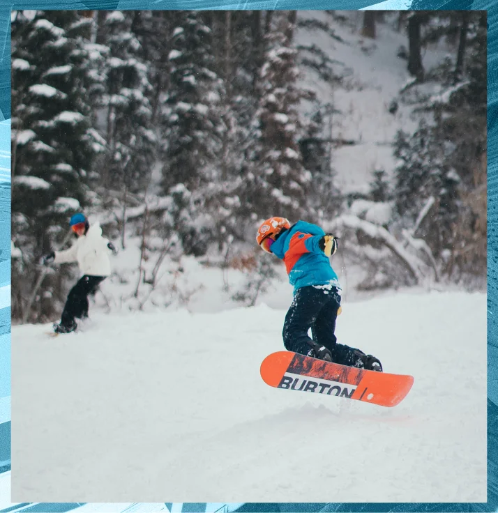 Two snowboarders are seen in action on a snowy slope surrounded by trees. The foreground snowboarder, using his S-Card, wears a blue and red jacket and is captured mid-turn on an orange Burton snowboard, while the other snowboarder is slightly blurred in the background.