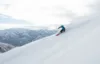 A skier dressed in blue and black is captured in mid-action, carving through fresh powder on a steep, snow-covered mountain slope with mountain peaks and a partly cloudy sky in the background. The precise technique showcased by the skier makes it look like an S-Card moment.