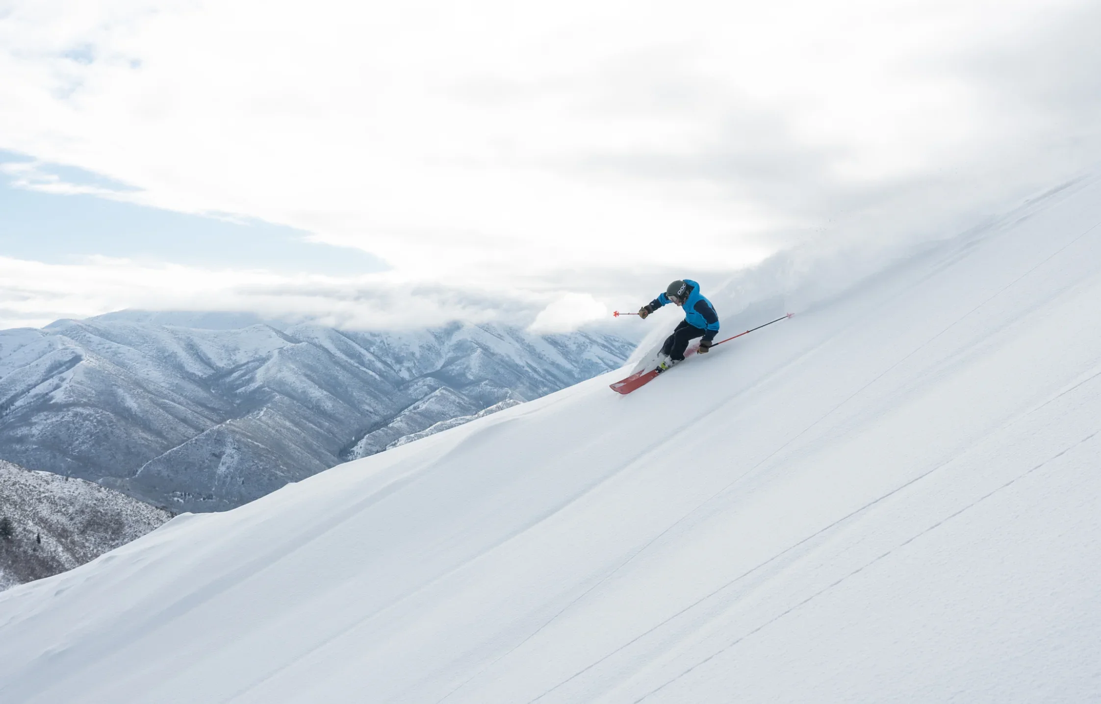 A skier dressed in blue and black is captured in mid-action, carving through fresh powder on a steep, snow-covered mountain slope with mountain peaks and a partly cloudy sky in the background. The precise technique showcased by the skier makes it look like an S-Card moment.