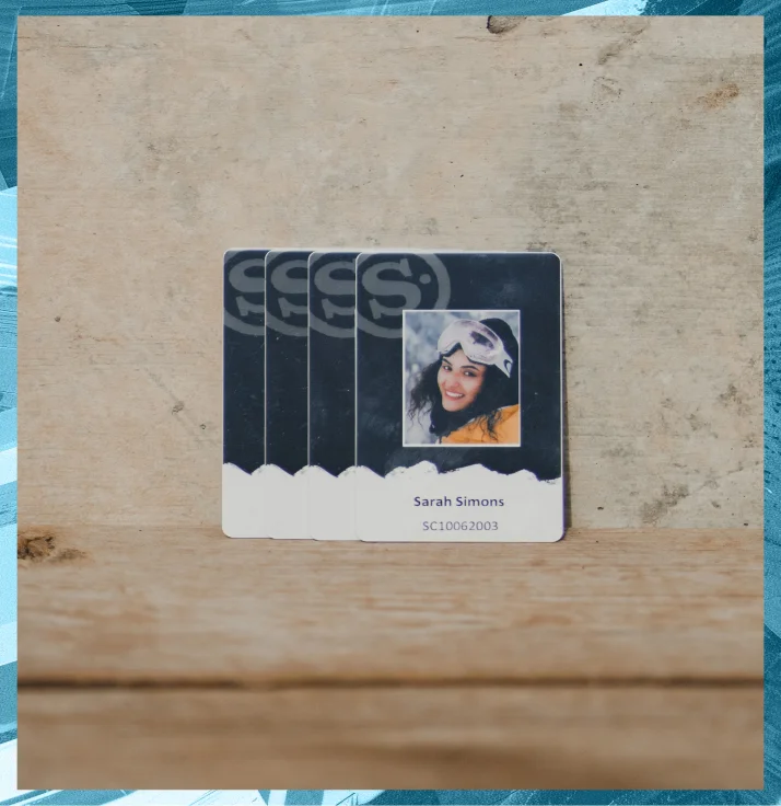 An S-Card displaying a photo of a woman in a white helmet and goggles, smiling. The card has the name “Sarah Simons” and the ID number “SC10062003” printed below the photo. The background is a textured surface with a blue and gray border.