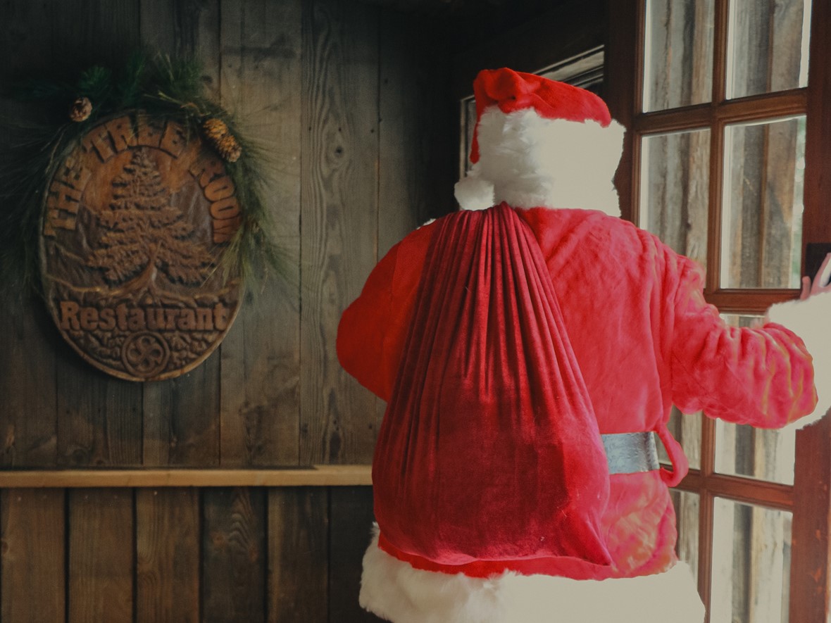 Santa Claus, wearing traditional red attire, stands by a wooden door with a sack over his shoulder. Beside him, a wooden plaque reads "The Tree Room Restaurant," adorned with festive greenery.