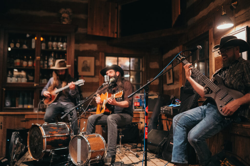 In a cozy, rustic room adorned for the holidays, a trio of musicians performs. The guitarist and banjo player wear festive hats, while the bassist plays alongside them. Instruments and a drum set create a warm, intimate musical setting perfect for the season's cheer.