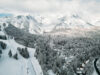 A scenic view of snow-covered mountains and forests. Ski slopes with trails can be seen alongside ski lifts, leading to small, scattered mountain lodges. Clouds partially obscure the mountain peaks, enhancing the pristine winter landscape.