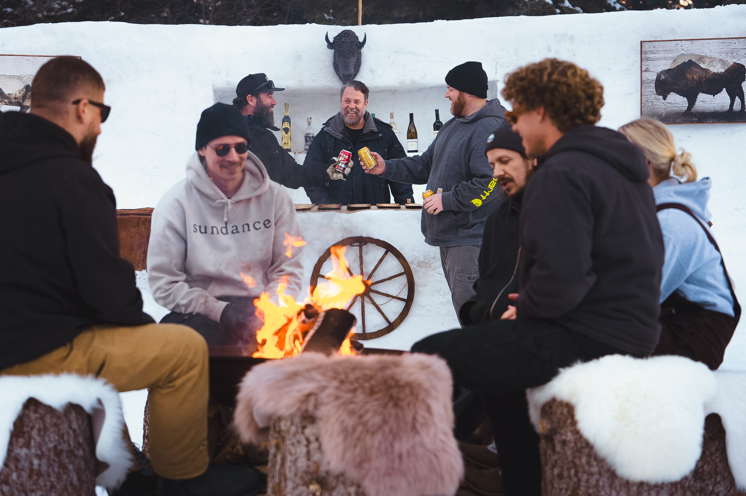 A group of people sit around a campfire on snow-covered seats with fur coverings. Behind them, a man serves drinks at an outdoor bar made of snow, decorated with a wagon wheel and a buffalo painting. It's a winter setting with casual attire.