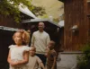 A group of people, including two adults and three children, are walking outdoors in front of rustic wooden buildings. The children are in the foreground, with greenery visible in the background.