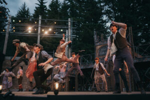 Performers in period costumes enthusiastically dance on an outdoor stage, surrounded by trees. A backdrop with scaffolding and a sign adds to the theatrical setting. Lighting illuminates the lively scene.