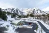 A winter scene of a ski resort with a green-roofed building surrounded by snow-covered mountains and trees. A road curves through the area, and a small crowd is visible near the entrance.