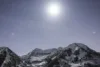 A snow-covered mountainous landscape under a starry sky. The moon shines brightly above, casting a soft glow on the peaks. The scene is serene and untouched, with stars scattered across the night sky.