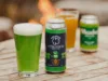 A can of Glitter Green Ale sits on a wooden table next to a glass of the same green beer. Two more glasses, one filled with amber liquid and another out-of-focus can, are in the background. A warm fire is slightly visible.