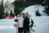 Two people wearing winter clothing stand side by side on a snowy slope. They hold snowboards and smile at the camera. Snow-covered trees and a ski lift are visible in the background.