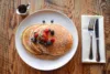 A delicious pancake breakfast awaits, with a plate of warm pancakes topped with strawberries, blueberries, and raspberries on a wooden table. A dusting of powdered sugar adds sweetness. Nearby, a small pitcher of syrup invites pouring, and silverware rests on a neatly folded napkin beside the plate.